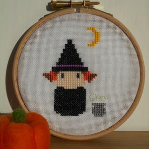 May include: A white embroidery hoop with a cross-stitch design of a witch with a black hat, orange hair, and a cauldron. The background is white with a yellow crescent moon.