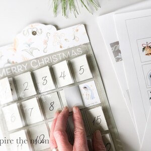 May include: A white and grey advent calendar with the words "Merry Christmas" printed on the top. The calendar has 24 numbered squares, each with a small window that can be opened to reveal a surprise. The calendar is decorated with a floral pattern.