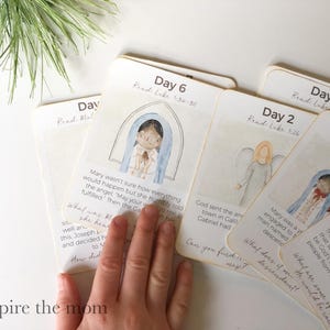 May include: A stack of illustrated cards with watercolor images and text. The cards are labeled with days and Bible verses, featuring images of angels and biblical figures. The cards are designed for educational purposes.