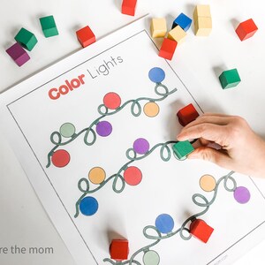 May include: A worksheet titled "Color Lights" with colorful light bulb illustrations. A hand places a green wooden block on the worksheet. Scattered around are other wooden blocks in various colors, including red, yellow, and purple.
