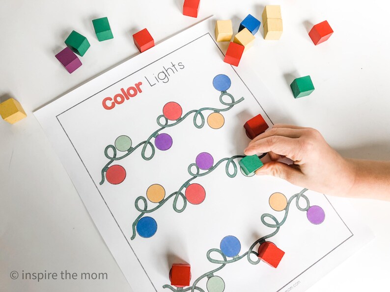 May include: A worksheet titled "Color Lights" with colorful light bulb illustrations. A hand places a green wooden block on the worksheet. Scattered around are other wooden blocks in various colors, including red, yellow, and purple.