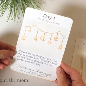 May include: A white card with the text "Day 1" and "Read: Isaiah 9:6". The card features a watercolor illustration of a mobile with stars, moons, and circles. The text below describes prophecies.