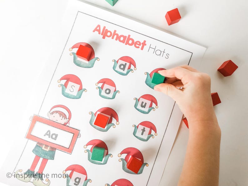 May include: A learning activity sheet titled "Alphabet Hats" with illustrations of elf hats and letters. A hand places a green block onto a hat. Red and green blocks are scattered around the sheet.