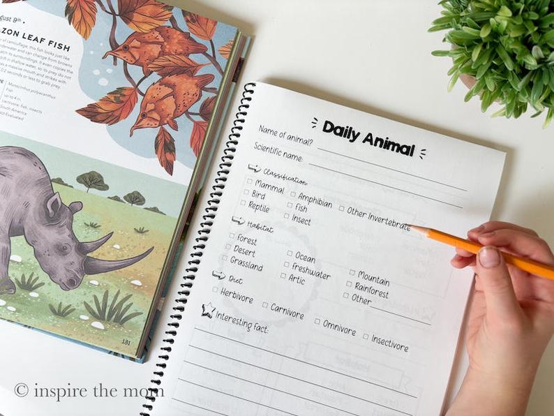 May include: A spiral-bound Daily Animal worksheet featuring sections for name, scientific name, classification, habitat, diet, and interesting facts, positioned next to an open illustrated book showing a rhinoceros and leaf fish.