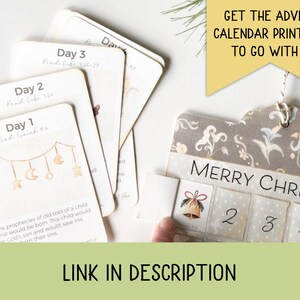 May include: Advent calendar with numbered days and a decorative design. The calendar has a "Merry Christmas" banner and includes cards with Bible verses. The image also includes the text "Get the Advent Calendar Printable to go with it" and "Link in Description."