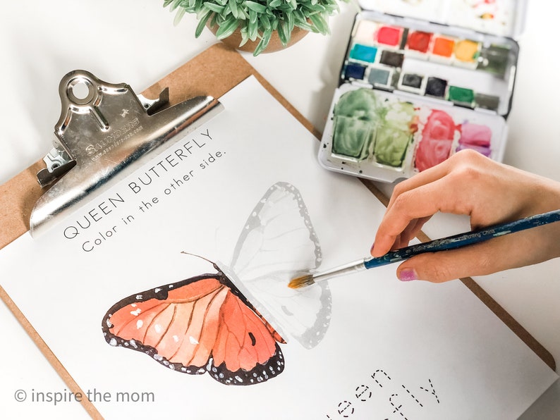 Pode incluir: Uma prancheta com uma ilustra&ccedil;&atilde;o de borboleta monarca para colorir. A borboleta est&aacute; parcialmente colorida em laranja e preto. Uma paleta de aquarela e um pincel tamb&eacute;m s&atilde;o vis&iacute;veis.