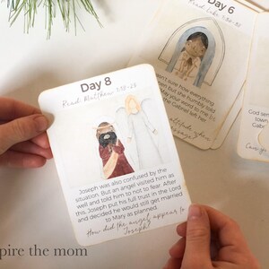 May include: A set of illustrated cards with watercolor images and text. One card, held by hands, features an angel and Joseph, with the text "Day 8" and "Read Matthew 1:18-25". Other cards are visible in the background.