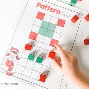 May include: A printed "Pattern Grid" worksheet with red and green squares, and a cartoon elf illustration. A hand places a red block on the grid. Several red and green blocks are scattered around the worksheet.