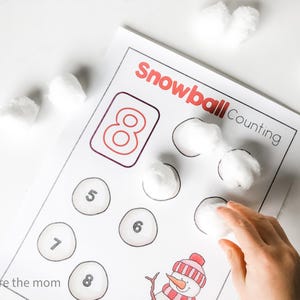 May include: A printed worksheet with the words "Snowball Counting" in red. The worksheet has circles with numbers and a drawing of a snowman. White cotton balls are placed on the worksheet, with a hand placing another cotton ball.