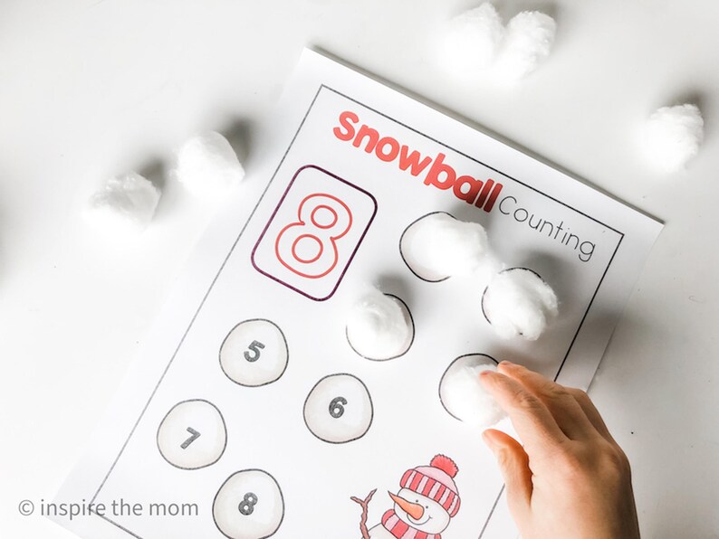May include: A printed worksheet with the words "Snowball Counting" in red. The worksheet has circles with numbers and a drawing of a snowman. White cotton balls are placed on the worksheet, with a hand placing another cotton ball.