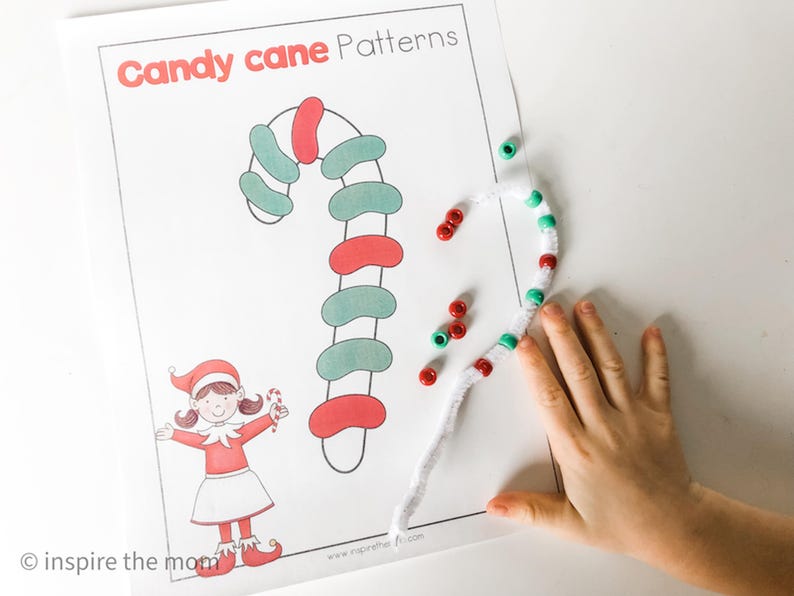 May include: A printed sheet with "Candy cane Patterns" in red text. A drawn candy cane is colored red and green. A child's hand is next to a pipe cleaner candy cane with red and green beads. A cartoon elf is also on the sheet.