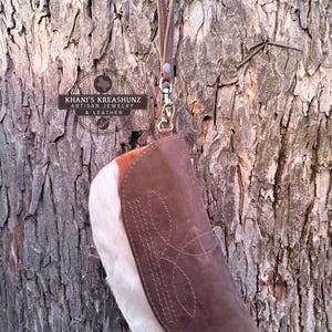 May include: A brown leather wristlet with a white and brown cowhide accent. The wristlet features a stitched design and a leather wrist strap with a silver-tone clasp. The background is a textured tree trunk.