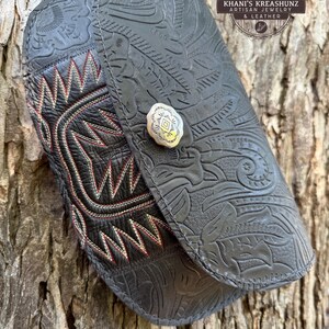 May include: A black leather wallet with intricate floral tooling and a silver button closure. The wallet features a decorative stitched design in red and white. The text "Khani's Kreashunz Artisan Jewelry & Leather" is visible.
