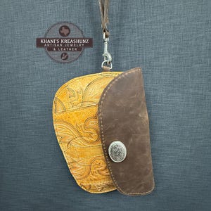 May include: A handcrafted leather wallet with a decorative pattern in shades of yellow and brown. The wallet features a dark brown flap secured by a silver button and a wrist strap. The brand name "Khani's Kreashunz Artisan Jewelry & Leather" is visible.
