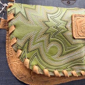 May include: Close-up of a handcrafted leather crossbody bag. The bag features a light green leather flap with intricate white and black stitching in a geometric pattern. The bottom and strap are tan leather with decorative stitching.