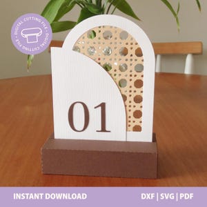 Pode incluir: Um sinal de número de mesa arqueado branco com o número 01 em castanho, sobre um padrão de palhinha bege. O sinal está montado numa base retangular castanha. Um emblema circular roxo no canto exibe o texto DIGITAL CUTTING FILE.