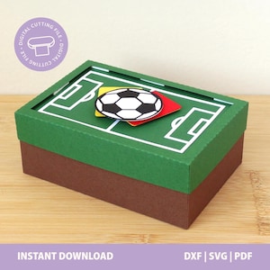 Pode incluir: Uma caixa de papel retangular com tampa verde apresentando um gráfico de campo de futebol branco, coberta com uma bola de futebol preta e branca, um cartão amarelo e um cartão vermelho. A base da caixa é marrom. Um logotipo circular diz DIGITAL CUTTING FILE.