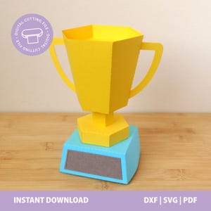 Puede incluir: Un trofeo de papel amarillo con dos asas, una base hexagonal y un pedestal azul claro con una placa rectangular gris. Una insignia circular morada en la esquina superior izquierda contiene el texto DIGITAL CUTTING FILE.