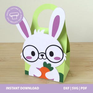 Pode incluir: Uma caixa de presente em forma de coelho de papel verde e branco, com uma alça. O coelho tem óculos redondos grandes, orelhas forradas de roxo e segura uma cenoura laranja. O texto na parte inferior diz "DOWNLOAD INSTANTÂNEO" e "DXF | SVG | PDF".