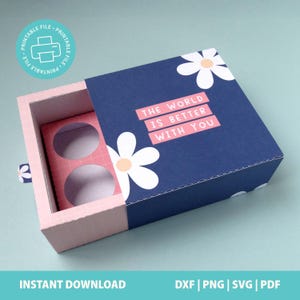May include: A rectangular sliding gift box with a dark blue outer sleeve featuring white daisy illustrations and the text THE WORLD IS BETTER WITH YOU in pink bars. The inner tray is light pink with a white grid pattern and two circular cutouts.