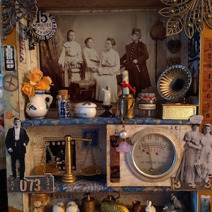 Op de afbeelding: Een vintage schaduwdoos met een sepia-foto, een miniatuur grammofoon en een manometer. De doos bevat diverse antieke items, decoratieve elementen en de tekst "No 12/258".