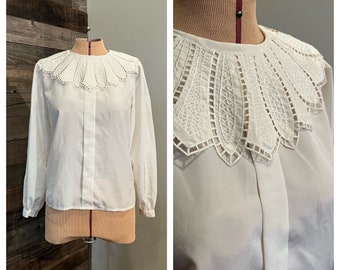 Puede incluir: Una blusa blanca de manga larga con un cuello de encaje festoneado y detallado. La blusa se abrocha por delante y tiene un estilo clásico y vintage. El cuello es el punto focal, añadiendo un toque de elegancia.