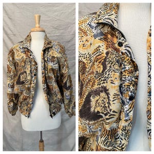 Puede incluir: Una chaqueta vintage con un estampado animal audaz que presenta tigres, leones y leopardos en tonos marrones, beige y negros. La chaqueta tiene cuello, cierre de botones y cremallera. La chaqueta se exhibe en un maniquí.