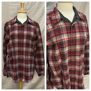 May include: A long-sleeve button-down shirt featuring a plaid pattern in shades of red, grey, and cream. The shirt has a pointed collar with a dark green inner lining, a front button placket, and long sleeves with buttoned cuffs on a mannequin.