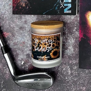 May include: A white candle with a black and white label that says "So very badass" and features a field of sunflowers. The candle has a wooden lid.