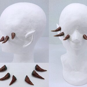 Puede incluir: Un conjunto de pequeños cuernos curvos de color marrón texturizado diseñados para aplicación facial. Los cuernos se muestran en una cabeza de maniquí blanca desde varios ángulos, exhibiendo su forma puntiaguda y su colocación.
