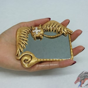 Puede incluir: Un espejo de mano rectangular dorado con detalles ornamentados. El espejo presenta un marco decorativo con un diseño alado y un emblema central. El espejo se sostiene en una mano, mostrando su tamaño y diseño.