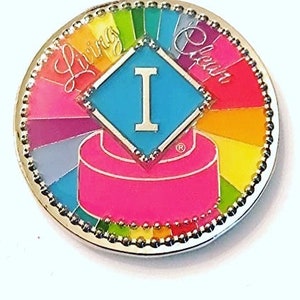 May include: A silver pin with a rainbow design and the words "Living Clean" around the edge. In the center is a blue diamond with a white "I" inside. The pin has a pink and white cake design at the bottom.