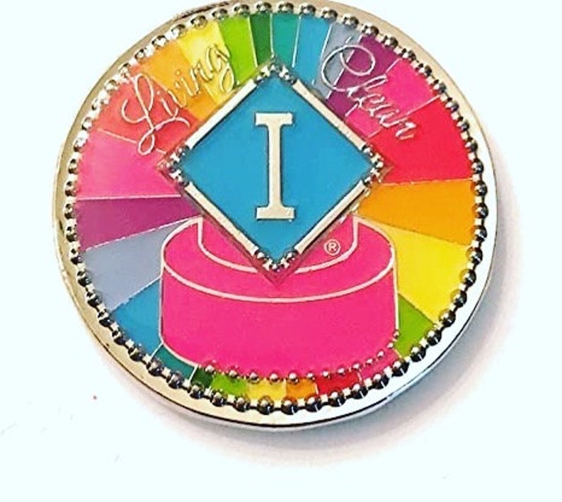 May include: A silver pin with a rainbow design and the words "Living Clean" around the edge. In the center is a blue diamond with a white "I" inside. The pin has a pink and white cake design at the bottom.
