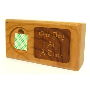 May include: A rectangular wooden block with a dark brown inset. The inset has the words "One Day At A Time" engraved in an elegant font. A circular indentation holds a square of green and white checkered adhesive.