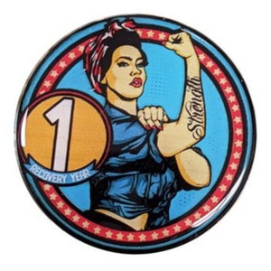 May include: A blue and red circular button with a woman in a blue shirt and red bandana with the words "Strength" tattooed on her arm. The woman is flexing her bicep. The button also has a yellow circle with the number "1" and the words "Recovery Year".