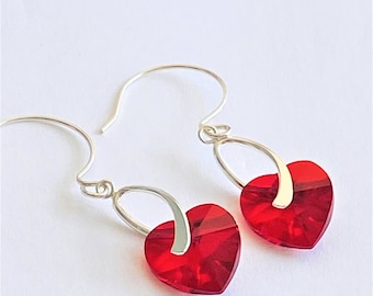 May include: A pair of heart-shaped earrings in a vibrant red colour. The earrings have a silver-coloured hook and a silver-coloured metal accent connecting the heart-shaped crystal to the hook. They are set against a white background.