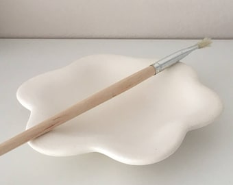 May include: A white ceramic dish shaped like a five-petaled flower. A wooden-handled paintbrush with a silver metal ferrule rests diagonally across the center of the dish. The dish has a smooth, matte finish and a shallow, concave interior surface.