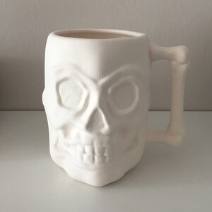 Könnte beinhalten: Weiße Keramiktasse in Schädelform mit Henkel. Die Tasse ist leer und bereit zum Bemalen.