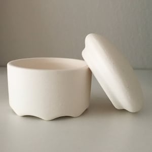 Puede incluir: Una caja de cerámica blanca para guardar pequeños objetos con tapa. La caja tiene una forma redondeada y un borde ligeramente ondulado. La tapa es ligeramente abovedada.