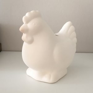 Puede incluir: Figurita de pollo de cerámica blanca con cuerpo redondeado y un pequeño pico. La figurita es un diseño simple sin detalles adicionales.