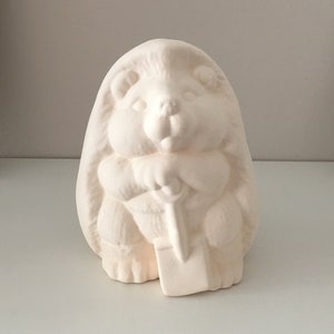Puede incluir: Una figurita de cerámica blanca de un erizo sosteniendo una pequeña pala. La figurita está sin pintar y lista para ser decorada.