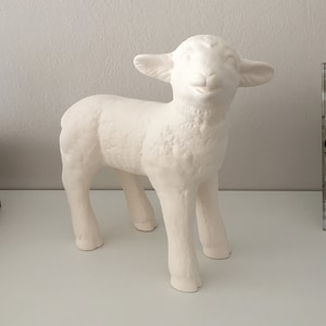 May include: White ceramic lamb figurine with a smiling face. The lamb is standing on all four legs and has a textured surface.