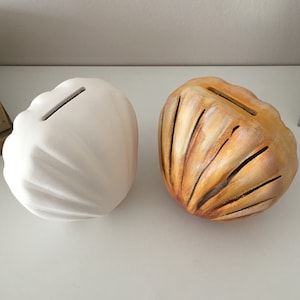 以下が含まれることがあります： 貝殻の形をしたセラミック製の貯金箱が2つ。1つは白で、もう1つは茶色と黄色のストライプで塗られています。