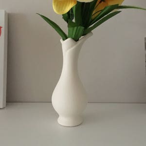 May include: A white ceramic vase with a unique, curved design holds a bouquet of yellow and green orchid flowers.