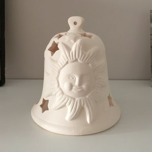 May include: White ceramic bell with a sun and stars design. The bell has cutouts in the shape of stars and a sun with a face.