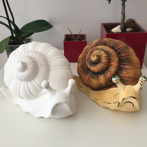 May include: Two ceramic snail sculptures. One is solid white, the other is painted with brown, yellow, and green details. The snails have detailed shell spirals and are placed on a white surface. Decorative garden art.