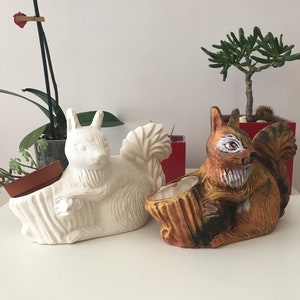 May include: Two ceramic squirrel planter pots. One is white with a brown tail and the other is brown with black and white details. Both are sitting on a table.
