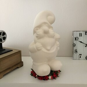 May include: A large, off-white ceramic gnome figurine with a pointed hat and beard. The gnome is standing on a bed of red berries and greenery. A vintage-style clock and a small decorative object are in the background.