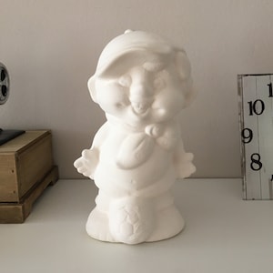 Puede incluir: Figurilla de cerámica blanca de un personaje de dibujos animados que lleva una gorra de béisbol y sostiene una bufanda. El personaje tiene una cara sonriente y está de pie sobre una base.