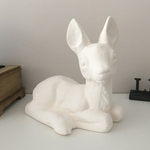 Puede incluir: Figurita de cerámica blanca de cervatillo en posición tumbada. El cervatillo tiene orejas grandes y una expresión dulce.